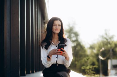 Beyaz bluzlu ve elinde telefonla dışarıda poz veren genç bir asistanın portresi. Yüksek kalite fotoğraf