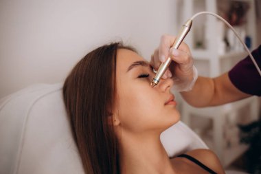 Kozmetik uzmanı, güzellik salonundaki güzel, genç bir kadının yüz derisinin Couperose tedavisini yapıyor. Kozmetoloji ve profesyonel cilt bakımı. Yüksek kalite fotoğraf