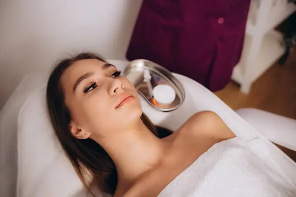 Professional Cosmetologist Cleansing Skin Of Young Asian Woman During Facial Beauty Treatments In Spa, Relaxed Korean Lady Enjoying Wellness Day At Beauty Parlor, Lying In Massage Table, Side View.