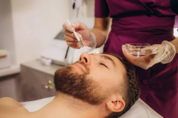 Beautician applying scrub onto young man\'s face in spa salon. High quality photo