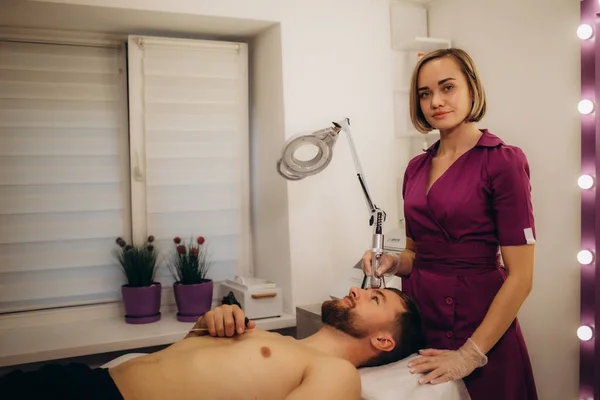 Professional female beautician performing face needle-free mesotherapy for male patient at cosmetology clinic. High quality photo