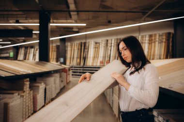  Kadın alıcı, kaplama örneklerini dikkatlice inceler. Kadın döşeme malzemelerini seçer. Kız ahşap malzemelerle vitrinin yanında duruyor. Satıcının konsültasyonunu bekliyor..