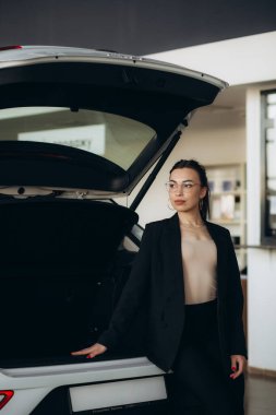 pretty young saleswoman in elegant suit dealer standing near car at new modern dealership. 