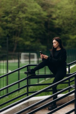Bir futbol sahasının arka planında telefon kullanan bayan sporcu.