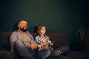 Mutlu baba ve küçük kız evde video oyunu oynuyorlar. Anaokulu kızı ve genç baba ellerinde av yastığı kontrolörleri, oturma odasında kanepede oturup aletlerle eğleniyorlar.