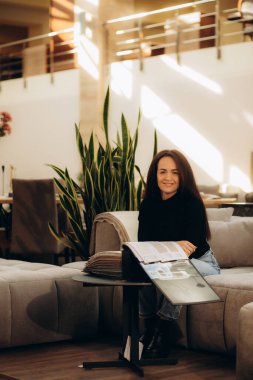 Sarı koltukta oturan ve elinde katalog tutan, sergi salonundaki yeni döşenmiş mobilyaları seçerken rüya gibi bakışlar atan hoş, çarpıcı, çekici, zarif, beyaz tenli, esmer kadın.