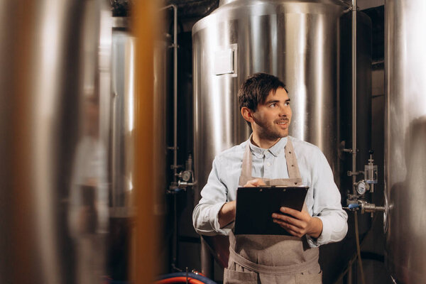Worker, brewer, business owner works on factory with equipment, modern tech. Satisfied smiling young attractive guy in an apron holds tablet and checks metal boilers at plant in brewery interior