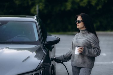 Küçük bir kamu istasyonundan şarj olan mutlu genç bir kadın, elektrikli arabanın yanında duruyor, kahve içiyor..