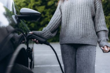 Caddedeki şarj istasyonunda şarj aletini mavi elektrikli arabasının prizine takan kadın.