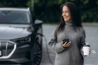 Kadın elektrikli arabasını şarj etmek için beklerken cep telefonuna bakıp kahve içiyor.