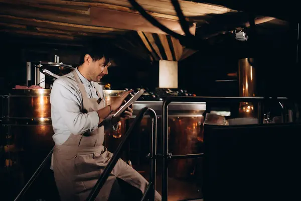 stock image Data collection and control or management of equipment at plant, small business and brewery. Millennial man worker in apron with tablet works with large boilers for fermentation of drink, free space