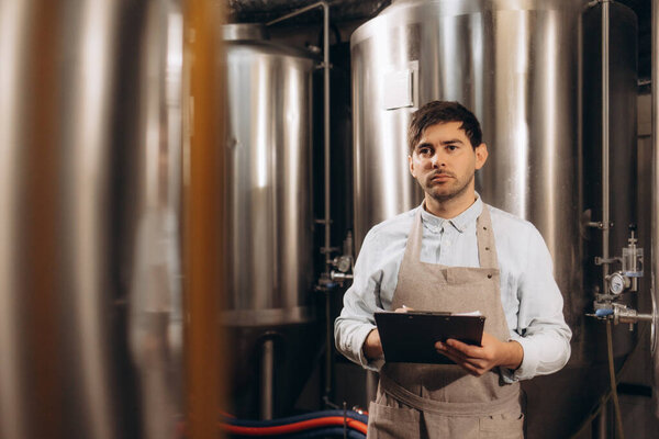 Data collection and control or management of equipment at plant, small business and brewery. Millennial man worker in apron with tablet works with large boilers for fermentation of drink, free space