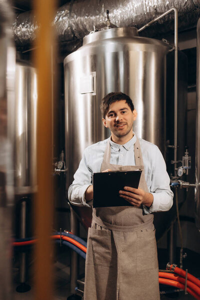 Brewer supervising the process of beer fermentation at the manufacturing with metal containers on the background