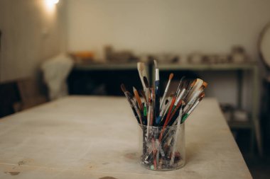 Artist supplies at wooden table. Paint brush in clay jug and art painter tools on wood desk background texture. Paintbrush for painting in artist studio workplace