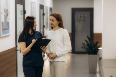 Gri üniformalı resepsiyonist ya da klinikçinin önünde tıbbi belgeleri imzalayan ya da dolduran genç bir bayan hasta.