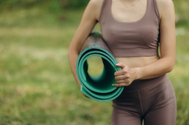 Kablosuz kulaklıklı, yoga minderiyle parkta kameraya gülümseyen mutlu beyaz kız. Sağlıklı yaşam tarzı kavramı