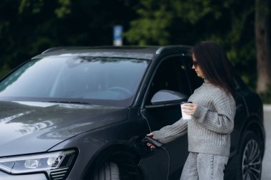 Kocaman gülümseyen genç bir kadın, uzaklara bakıyor, küçük bir kamu istasyonundan araba aküsünü şarj ediyor, elektrikli arabanın yanında duruyor, kahve içiyor ve akıllı telefondan konuşuyor.