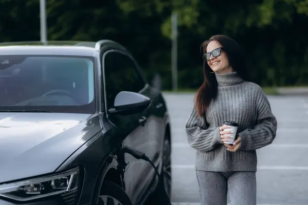 Stock image Portrait of a young woman standing near her electric car charging on a public station. Concept of modern lifestyle and success