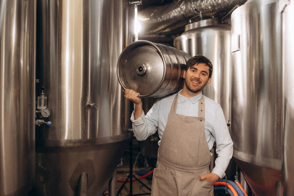 Portrait of male brewer carrying metal container at brewery factory. Young man holding a keg on shoulder at warehouse.