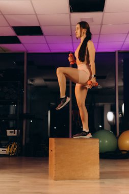 Fit millennial woman using wooden cubes as props in her fitness routine in a gym or a health club, performing a strength and conditioning workout to improve cardiovascular endurance or maintain weight