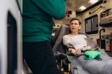 Yaralı hastayla ilgilenen sağlık görevlileri iğne yapıyor. Genç erkek sağlık görevlileri serum solüsyonu tutuyor. Kadın doktor, hastanın kalbini dinliyor.