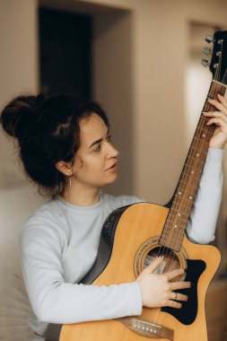 Gülümseyen ve gitar çalmayı öğrenen esmer, çekici bir kadın ev içinde rahat bir şekilde oturuyor. Genç mutlu kadın müzik aleti performansıyla hobisinin keyfini çıkarıyor hasır sandalyede dinleniyor.