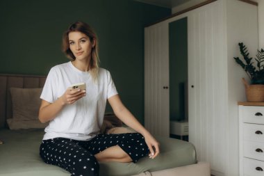 Elinde telefon olan pijamalı bir kadın yatağa oturur.