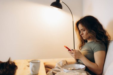 Bir kadın kahvaltı yapar ve kanepede telefonuna bakar.