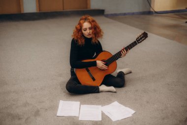 Güzel sarışın kızın tam vücut fotoğrafı. Oturup gitar çal. Çizgili giysiler giy.