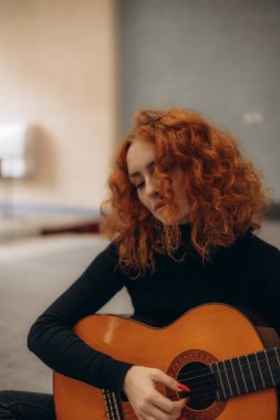 Mutlu genç bir kadın, hafta sonları evdeki rahat oturma odasında oturup dinlenirken gitar çalıyor..