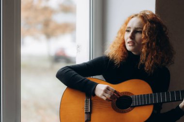 Kızıl saçlı bir kız pencerede gitar çalıyor.