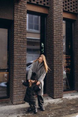 Bir anne ve kızı, sıcacık giyimli, evlerin önündeki kış caddelerinde yürürler.