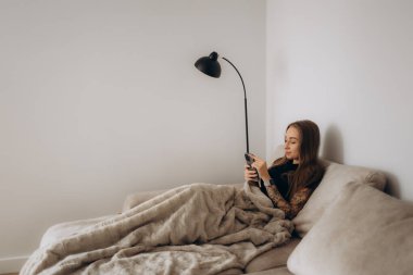Pijamalı, gülümseyen ve koltukta otururken cep telefonu kullanan mutlu bir kadın resmi.