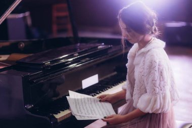 Şık giyinen genç bir kadın, müzik notaları üzerinde çalışırken, zarif bir piyanonun başında, zarif ve sakin bir atmosfer yaratıyor, müzik çalışması ya da performans hazırlığı için sıcak ortam aydınlatması kuruyor..