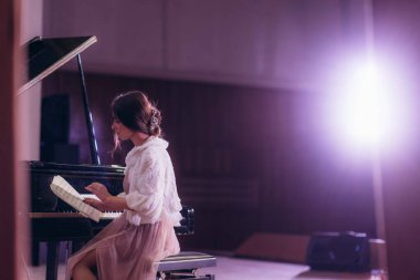 Genç bir kadın, yumuşak, kasvetli bir aydınlatma ortamında piyanonun başında oturmuş, yaratıcılık ve sanatsal özveriyi çağrıştıran notalara odaklanır..