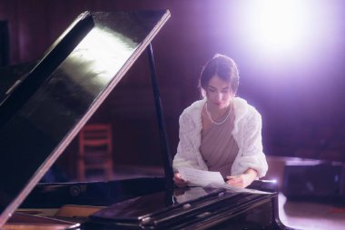 Resmi bir elbise ve beyaz bir sargı giymiş sofistike bir kadın, büyük bir piyanonun yanında müzik örtüsü tutuyor, loş bir performans ortamında, çarpıcı bir ışık efektiyle aydınlatılıyor..