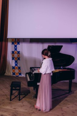 A woman elegantly dressed in a white fur shawl and pink skirt stands beside a grand piano, preparing for a performance on a dimly lit concert stage. clipart