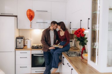 Modern bir mutfakta, dekorasyonlar, güller ve balonlarla çevrili romantik bir anı paylaşan bir çift, sevgi dolu ve samimi bir ortam yaratıyor..