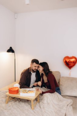 Bir çift, evdeki ahşap tepside suşinin tadını çıkarırken samimi bir anı paylaşıyor. Kalp şeklinde bir balon ve sıcak ortam aydınlatmasıyla geliştirilmiş sevgi ve bağları ifade ediyor..
