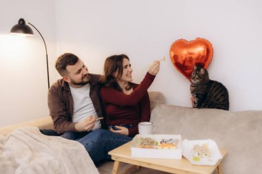 Sevgi dolu bir çift, kedileri ve dekoratif kırmızı kalp şeklinde bir balonla birlikte sıcak bir oturma odasında sushi yiyerek bir an paylaşır..