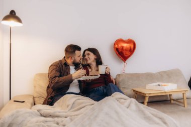 Çift evde samimi, samimi bir anı paylaşıyor, kalp şeklinde bir balonla çevrili, battaniyeyle kaplı bir kanepede dinleniyor, ve sıcak bir gülümseme paylaşıyor..