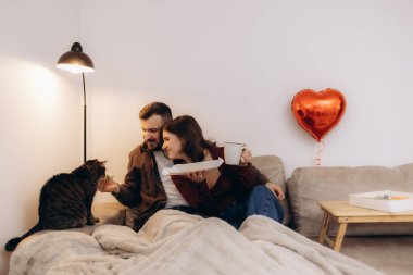Romantik bir çift, kanepede kahve ve atıştırmalıklarla rahatlar, yanında da bir kedi olur..
