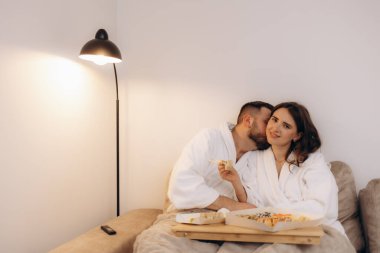 Beyaz bornozlu bir çift evde dinleniyor, birlikte suşinin tadını çıkarıyorlar. Sıcak, samimi bir ortamda rahat bir şekilde otururlar, bir yemeği ve bir bağı paylaşırlar..