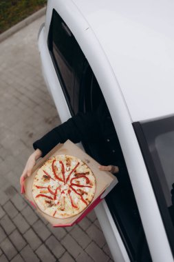 Bir insan sıcak, taze hazırlanmış bir pizza kutusunu tutarak bir arabanın camından kolunu uzatıyor, hızlı ve uygun bir yemek servisi sergiliyor ve taze ve lezzetli yiyeceklerin cazibesini vurguluyor..