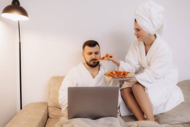 Çift kanepede oturuyor, bornoz giyiyor, pizzanın tadını çıkarıyor ve rahat bir ev ortamında dizüstü bilgisayarla çalışıyor..