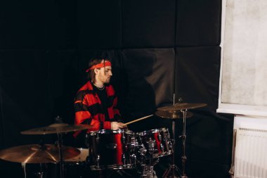 A young man wearing a vivid outfit plays drums in a studio with acoustic panel walls. Captures the passion of a music artist rehearsing in a professional practice environment. clipart