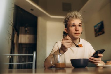 Genç bir yetişkin, bir elinde cep telefonu, diğer elinde çatal bıçak, iyi ışıklandırılmış bir iç mekanda kahvaltıdan zevk alıyor. Ayar rahat ve rahat bir atmosferi teşvik ediyor..