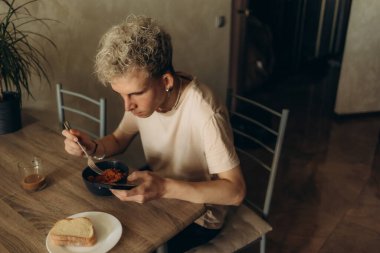 Kıvırcık saçlı genç bir adam ahşap bir masada oturur ve sıcak bir ev ortamında kahvaltı yapar. Masada bir tabak ekmek ve bir bardak kahve var..