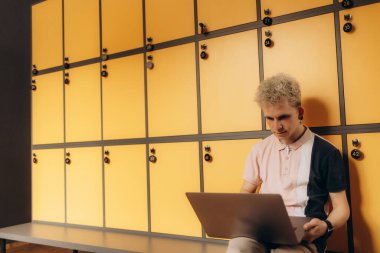 Modern sahnede, canlı sarı dolapların yanında dizüstü bilgisayarı olan bir adam görülüyor. Görüntü, gençlik, teknoloji ve eğitim veya kişisel çalışma alanı temaları için uygun bir gündelik ortamı vurguluyor.