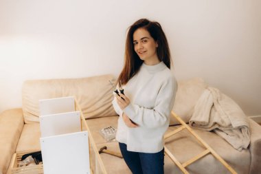 A woman is confidently assembling furniture in her cozy living room, holding a screwdriver. This scene represents do-it-yourself efforts, creativity, and home improvement within a comfortable, welcoming environment. clipart